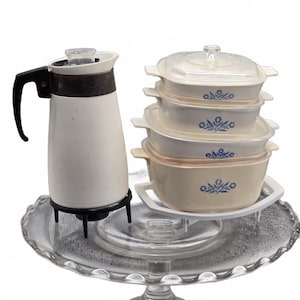 Può includere: Una caffettiera bianca con manico nero e una pila di quattro pirofile rettangolari in vetro bianco con un motivo floreale blu. Le pirofile hanno coperchi in vetro trasparente e sono disposte su un'alzata per torte in vetro.