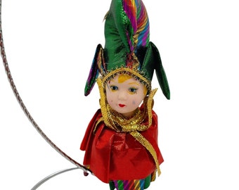 May include: A porcelain doll ornament with a jester hat. The doll is wearing a red and green costume with gold trim. The hat is green with rainbow stripes and gold trim.