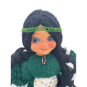 May include: A yarn doll with long black braids, a green dress, and a beaded necklace. The doll has a smiling face with blue eyes and a green beaded headband.