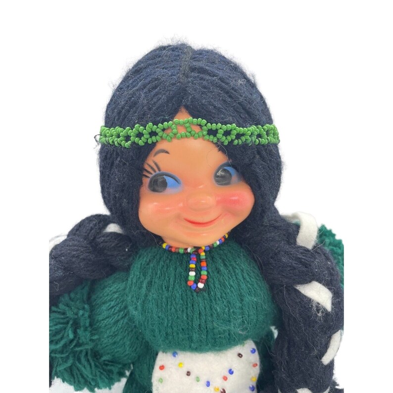 May include: A yarn doll with long black braids, a green dress, and a beaded necklace. The doll has a smiling face with blue eyes and a green beaded headband.