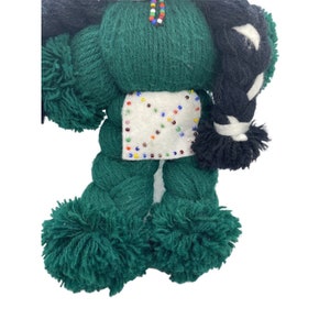 May include: A green yarn doll with black yarn hair, white yarn clothing, and colorful beads. The doll has pom-poms for feet and hands.