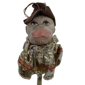 May include: A whimsical monkey ornament with a brown felt hat and a white and gold ruffled collar. The monkey has a gray face with brown eyes and a pink nose.