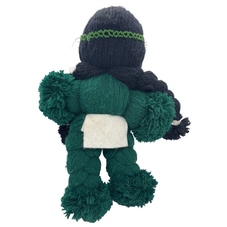 May include: A green yarn doll with black yarn hair and a white fabric apron. The doll has pom-poms for arms and legs.
