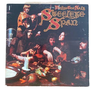 以下が含まれることがあります： Steeleye Spanの「Below the Salt」のアルバムカバー。カバーは、装飾的な赤い文字でバンド名が書かれた暗い背景が特徴です。食べ物とろうそくの火が灯ったテーブルを囲む人々のグループ。