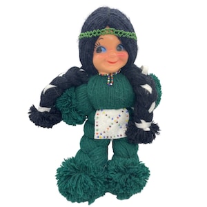 May include: A green yarn doll with black yarn hair, a beaded necklace, and a white beaded skirt. The doll has a smiling face with blue eyes.
