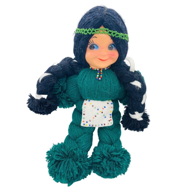 May include: A handmade yarn doll with long black braids, a green dress, and a white beaded apron. The doll has a smiling face with blue eyes and a green headband.