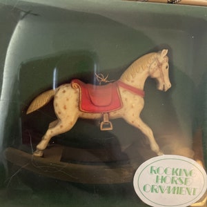 May include: A white rocking horse ornament with brown and gold accents. The horse has a red saddle and a gold bridle. The ornament is on a wooden base and has a gold string for hanging. The text "ROCKING HORSE ORNAMENT" is printed on a white oval label.