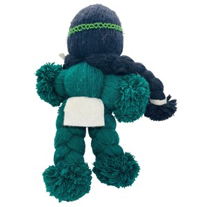 May include: A handmade yarn doll with a green body, black hair, and a white fabric patch. The doll has pom-poms for arms and legs.