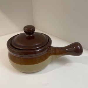 May include: A small brown and beige ceramic pot with a lid and a handle. The pot is glazed and has a rustic look.