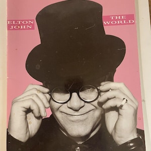 May include: A black and white photo of Elton John wearing a black top hat, glasses, and a black shirt. The background is pink with the text "Elton John" and "The World" in white.