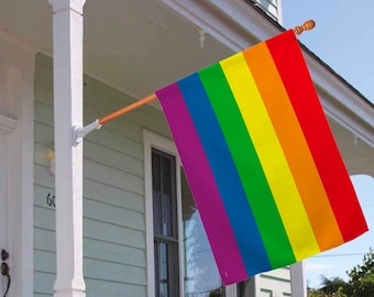 May include: A rainbow pride flag is displayed on a house with a white porch and green siding. The flag is attached to a wooden pole and is flying in the wind.