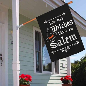 May include: Black Halloween flag with the text "Not all Witches Live in Salem" in white script. The flag features a witch hat with an orange band. The flag is hanging on a porch with red flowers in pots.