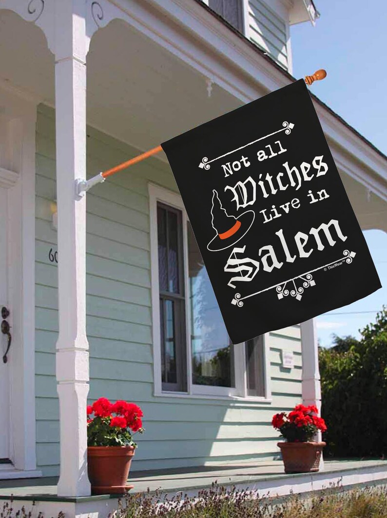 May include: Black Halloween flag with the text "Not all Witches Live in Salem" in white script. The flag features a witch hat with an orange band. The flag is hanging on a porch with red flowers in pots.