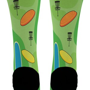 May include: A pair of green socks featuring a disc golf course design. The socks have a light green background with illustrations of disc golf baskets, orange and yellow discs, and blue and green course elements. The heels and toes are black.