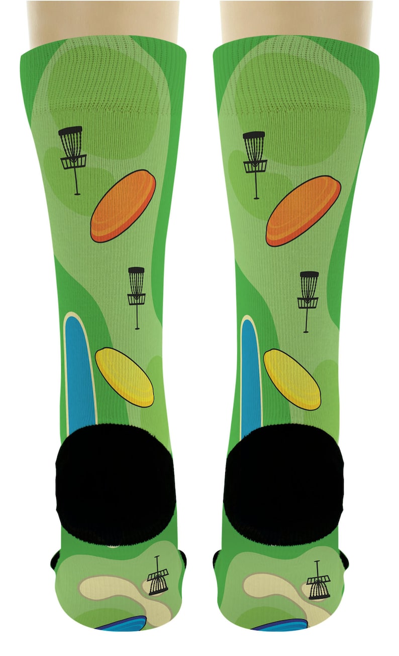 May include: A pair of green socks featuring a disc golf course design. The socks have a light green background with illustrations of disc golf baskets, orange and yellow discs, and blue and green course elements. The heels and toes are black.