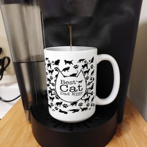 May include: A white ceramic mug with a black handle and the text "But First, Coffee" in a bold, black font. The mug is set against a blurred background, highlighting its simple design.