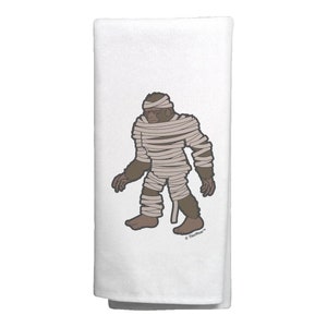 May include: A white kitchen towel with a brown cartoon illustration of a Bigfoot mummy. The mummy is wrapped in white bandages and has red eyes. The text "ThisWear™" is printed in the bottom right corner of the towel.