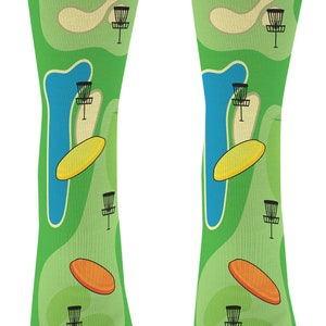 May include: Green socks with a disc golf course design. The socks feature a repeating pattern of disc golf baskets, discs, and water hazards. The socks have black toes and heels.