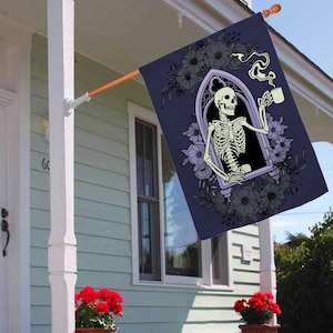 May include: A purple and black garden flag with a skeleton holding a steaming cup of coffee. The skeleton is sitting in a window with floral details.
