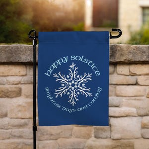 May include: A navy blue garden flag with the words "happy solstice" and "Brighter Days are coming" encircling a white snowflake design. The flag is hanging on a black metal pole in front of a stone wall.