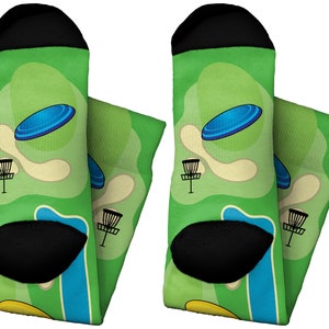 May include: A pair of green socks featuring a disc golf course design. The socks have a black toe and heel, with a pattern of a blue disc, a basket, and a course layout in shades of green, beige, and blue. The socks are designed for comfort and style.