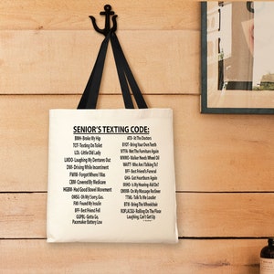 May include: A cream-colored tote bag with black handles hangs from a black anchor-shaped hook. The bag features the text "SENIOR'S TEXTING CODE:" followed by a list of abbreviations and their meanings. The bag is hanging on a wooden wall.