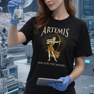 May include: Black t-shirt featuring the word "ARTEMIS" above an illustration of a woman with a bow and arrow. The text "AIM FOR THE MOON" is below the illustration. The shirt is made of a soft material.
