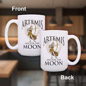 May include: Two white ceramic mugs with the word "ARTEMIS" in black script, and a graphic of a woman with a bow and arrow. The text "AIM FOR THE MOON" is below the graphic. The mugs have large, curved handles.