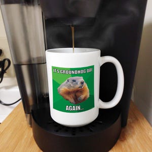 May include: White coffee mug with a green, white, and black image of a groundhog. The text on the mug reads "It's Groundhog Day Again..."
