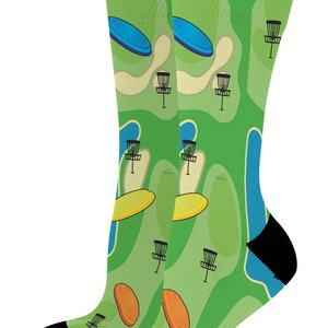 May include: A pair of knee-high socks featuring a vibrant green disc golf course design. The socks display a repeating pattern of disc golf baskets, discs, and course elements in shades of green, blue, yellow, and orange, with black heels and toes.