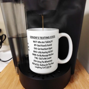 May include: White coffee mug with black text that reads "Senior's Texting Code:" followed by a list of abbreviations and their meanings. The mug is being filled with coffee from a coffee maker.