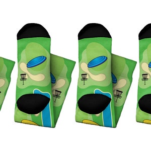 May include: Four pairs of socks with a green background featuring a blue disc flying over a disc golf course. The socks have black heels and toes, and the design includes a basket and a tee area.