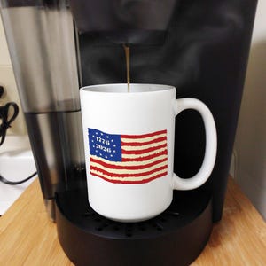 May include: A white ceramic mug with a patriotic American flag design, including the years 1776 and 2026. The mug is positioned in a coffee maker, with coffee being poured into it. The flag features red and white stripes and a blue field with white stars.