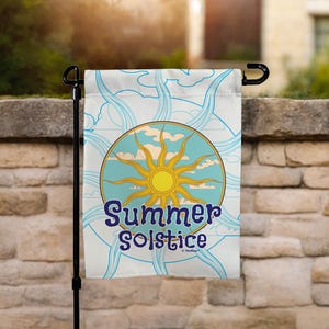 May include: A white garden flag with a circular design featuring a yellow sun, blue sky, and clouds. The words "Summer Solstice" are written in blue script. The flag is hanging on a black metal pole.