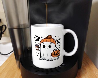 May include: A white ceramic coffee mug with a cartoon ghost wearing an orange and white hat and holding a pumpkin. The ghost is holding a small cup of coffee. The mug has a black handle and is filled with coffee.