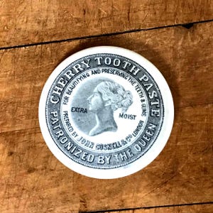 May include: A round, white container with the text "CHERRY TOOTH PASTE" in a circular design. The label features a profile of a woman and the words "EXTRA" and "MOIST". The container is "PATRONIZED BY THE QUEEN".