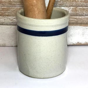 May include: A speckled, off-white ceramic kitchen utensil holder with a dark blue band. Two wooden kitchen tools are inside the holder. The holder has a cylindrical shape and a slightly flared rim. The background is a white surface.