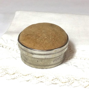 May include: A round, antique pin cushion with a light brown, velvet-like top. The cushion sits in a silver-colored metal base with a decorative, dotted pattern around the sides. The background is a white, textured fabric.