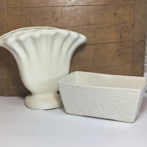 May include: A cream-colored ceramic vase with a wide, scalloped, fan-like top and a pedestal base, positioned next to a rectangular cream-colored ceramic planter featuring a subtle, embossed floral pattern on its exterior surface.