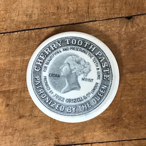 May include: A round, white Cherry Tooth Paste tin lid with black text and a gray illustration of a woman's profile. The text includes "Extra Moist" and "Patronized by the Queen."
