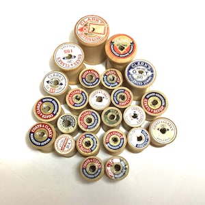 May include: A collection of vintage wooden sewing thread spools arranged in a pyramid shape. Each spool features a circular paper label with brand names like Clark's, Coats & Clark's, and Star, along with various color schemes and text.