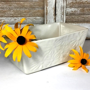 May include: A rectangular off-white ceramic planter featuring a subtle embossed floral pattern on its exterior surface. The planter is decorated with bright yellow black-eyed Susan flowers with dark brown centers placed around the container.