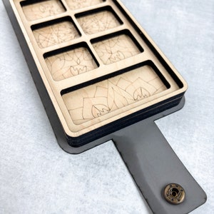 May include: A wooden box with a black trim and a gray leather strap. The box has eight compartments, each with a different laser-etched design. The box has a brass button on the strap.