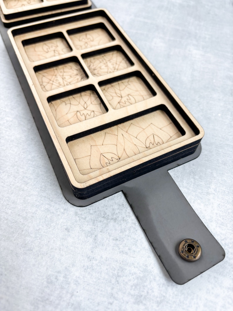 May include: A wooden box with a black trim and a gray leather strap. The box has eight compartments, each with a different laser-etched design. The box has a brass button on the strap.