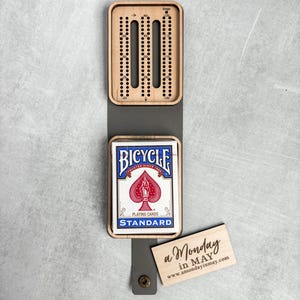 以下が含まれることがあります： Bicycleのトランプと「a Monday in MAY www.amondayinmay.com」と書かれた小さな木製の看板が付いた木製クリベッジボードです。