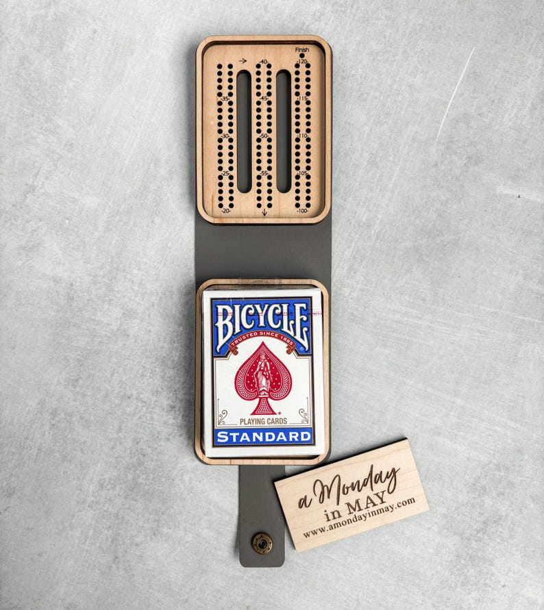 以下が含まれることがあります： Bicycleのトランプと「a Monday in MAY www.amondayinmay.com」と書かれた小さな木製の看板が付いた木製クリベッジボードです。