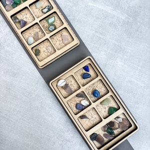 May include: A wooden tray with two sections, each with nine compartments, for storing small items. The tray is gray and has a leather strap. The compartments are filled with colorful stones.