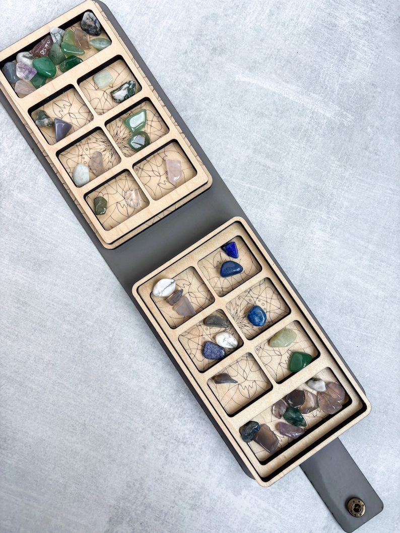 May include: A wooden tray with two sections, each with nine compartments, for storing small items. The tray is gray and has a leather strap. The compartments are filled with colorful stones.