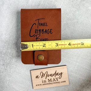 以下が含まれることがあります： 前面に「TRAVEL CRIBBAGE BOARD」と刻まれた茶色の革製の旅行用クリベッジボード。ボードは黄色のメジャーで測定されており、幅は約7.6cmであることがわかります。手前に「a Monday in MAY www.amondayinmay.com」と書かれた小さな木製の看板があります。