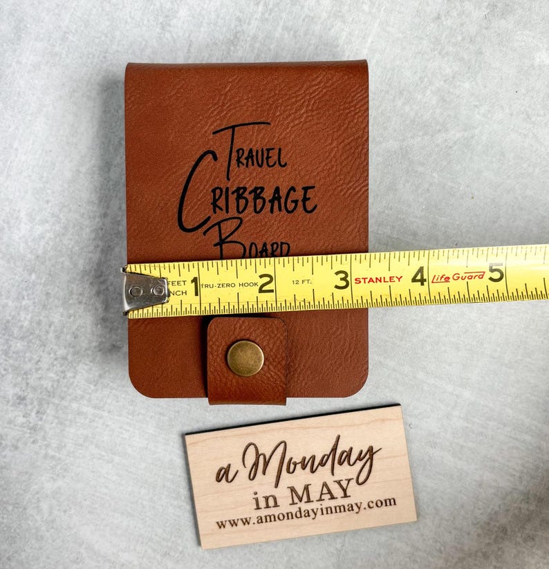 以下が含まれることがあります： 前面に「TRAVEL CRIBBAGE BOARD」と刻まれた茶色の革製の旅行用クリベッジボード。ボードは黄色のメジャーで測定されており、幅は約7.6cmであることがわかります。手前に「a Monday in MAY www.amondayinmay.com」と書かれた小さな木製の看板があります。